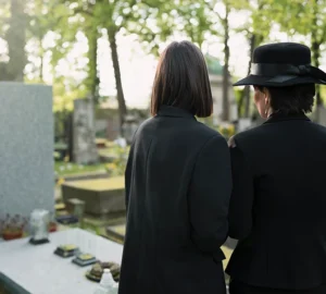 Two people wearing black, standing by a gravesite with headstones in the background.
