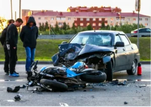 A motorcycle laying on the ground and a car with severe front-end damage, while two people on the side of a road. 