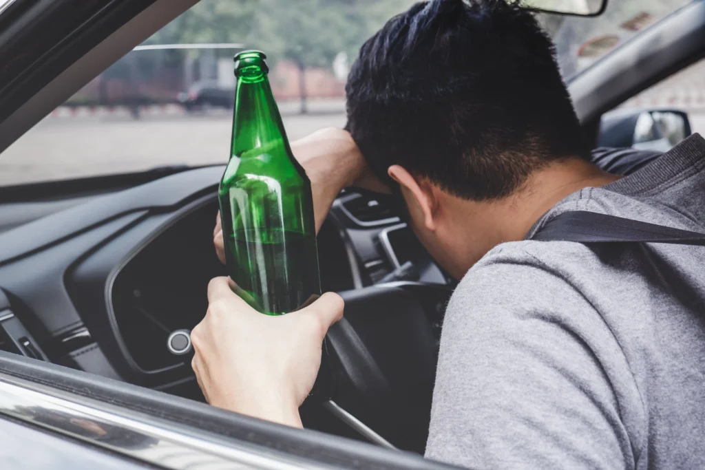 A person driving while holding a green beer bottle.